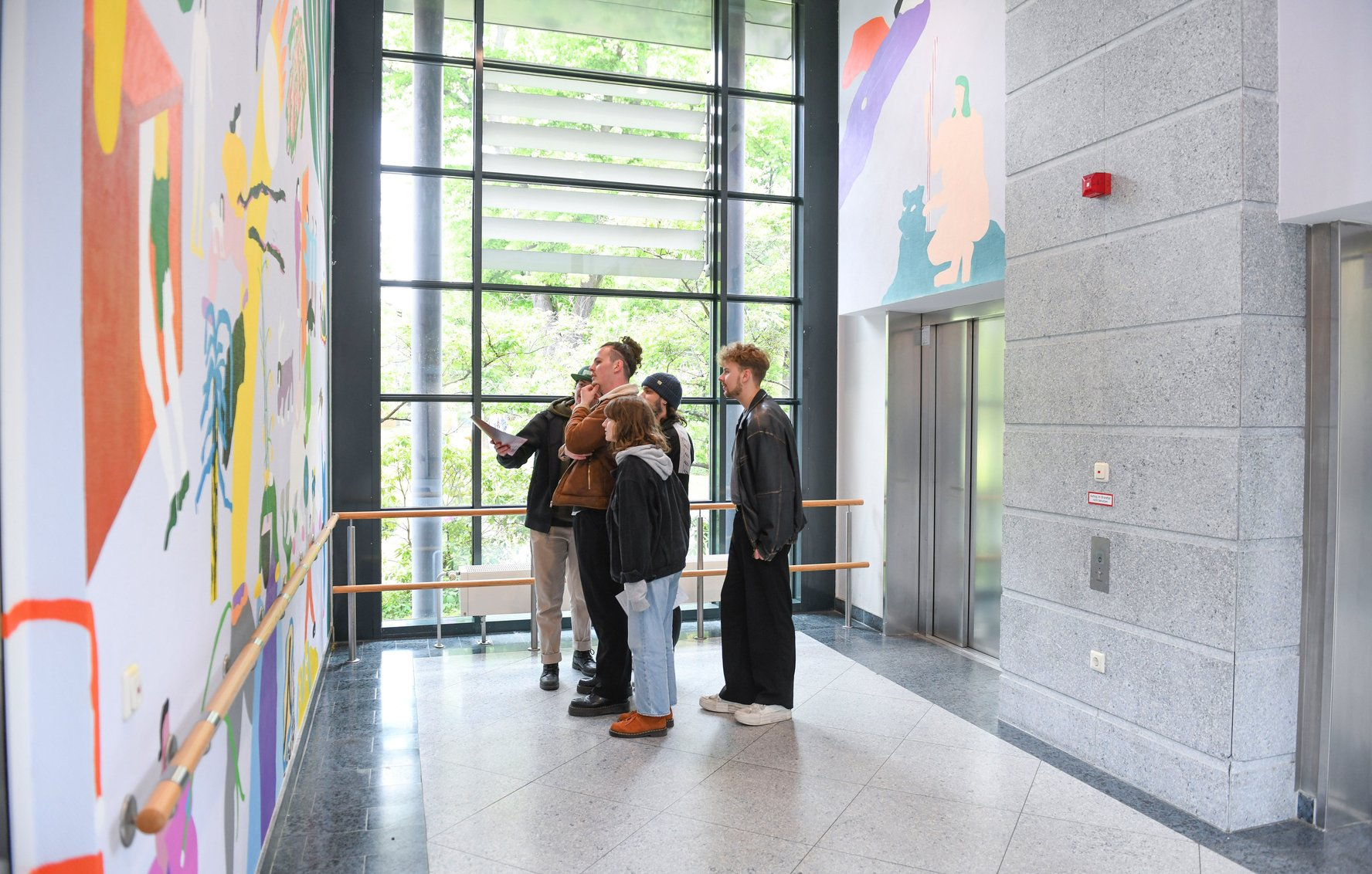 Eine Gruppe junger Menschen steht in einem hellen Foyer mit großer Fensterfront und betrachtet ein farbenfrohes Wandgemälde.