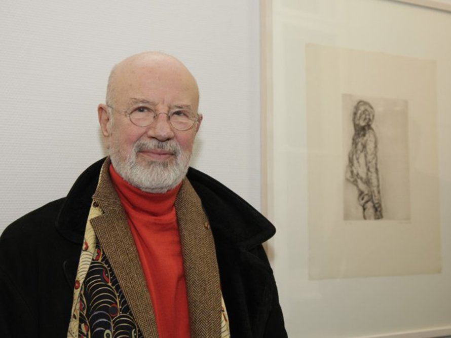 Künstler Marwan mit weißem Bart, Brille und rotem Rollkragenpullover steht neben einer seiner Radierungen mit Figurenmotiv.